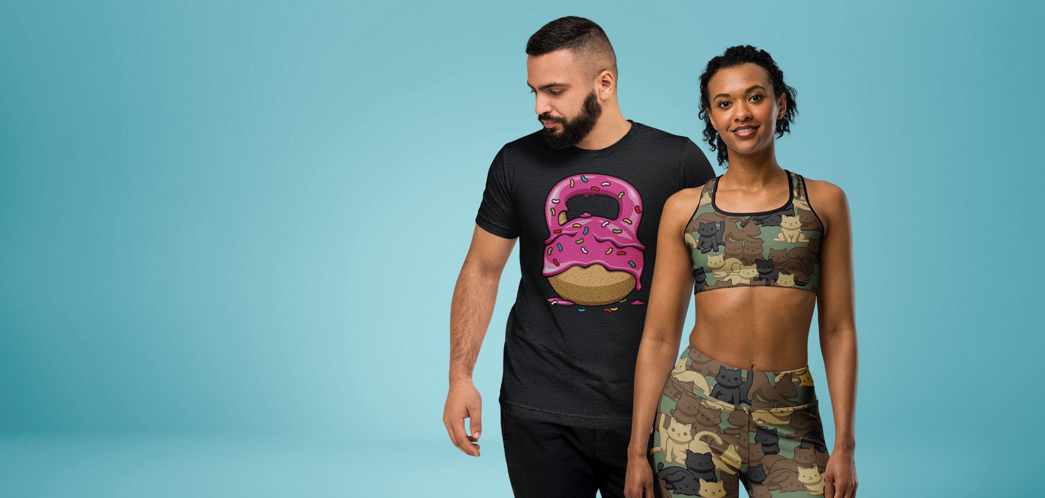 A man and a woman modeling activewear against a solid teal-blue studio background. The man wears a black t-shirt with a pink and brown kettlebell donut graphic, and the woman wears a matching cat camouflage-patterned sports bra and leggings set.